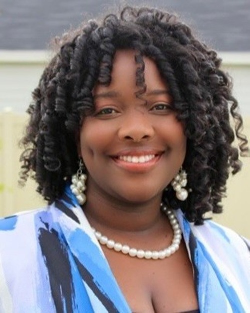 Kyra Lucas-Millbrook A woman with dark curly hair is wearing a blue top and pearls and smiling at the camera