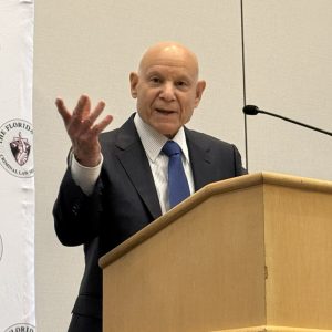 A man wearing business attire is lecturing at a podium with one hand outstretched